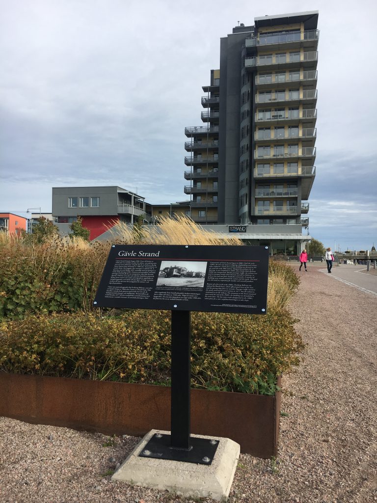 Kulturskylt märkt Gävle strand intill plantering vid grått höghus