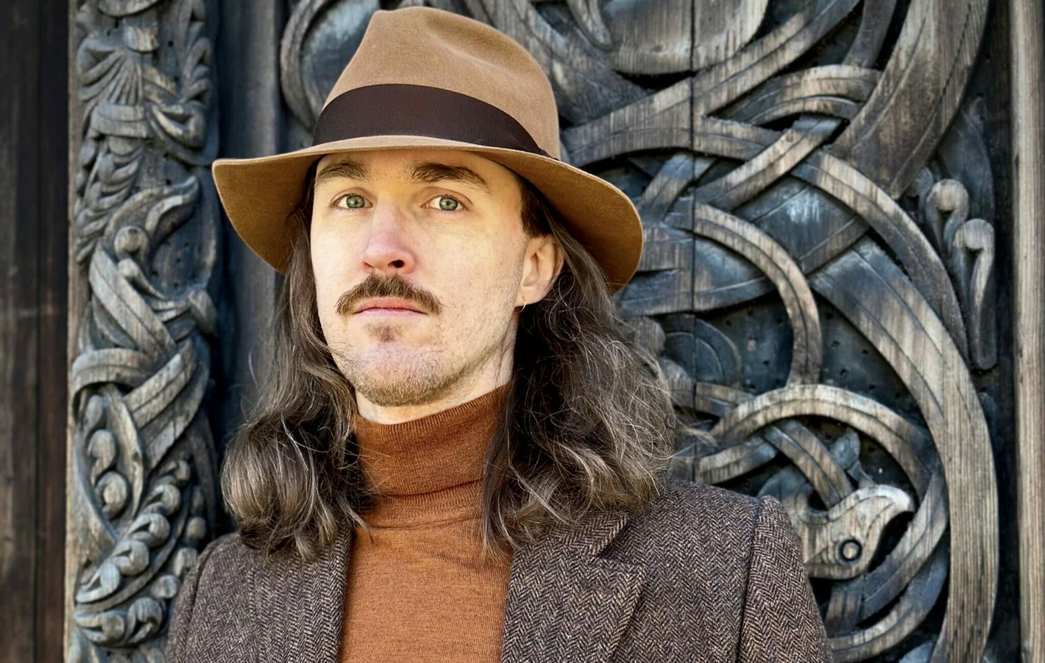 En man i en brun hatt, med långt brunt hår och en polotröja och kavaj.