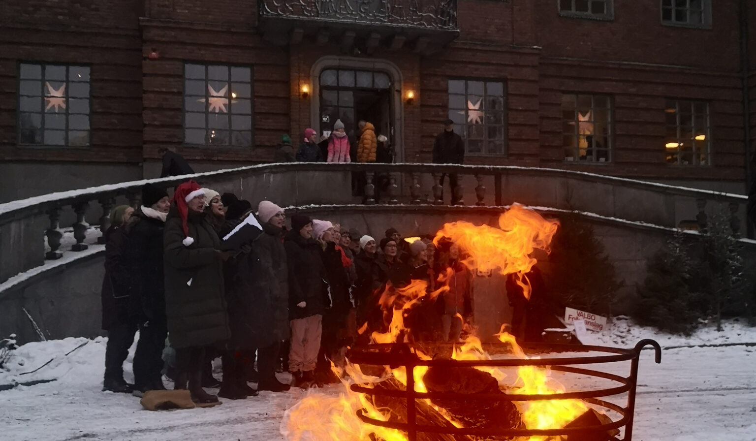 En kör står och sjunger utanför Länsmuseet Gävleborg. Snö på marken. En vedkorg står i förgrunden till bilden. Bilden är tagen på julmarknaden 2022
