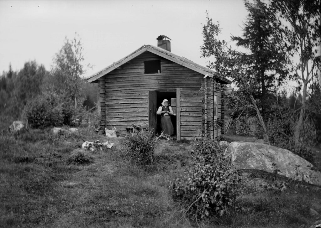 Ett svartvitt foto med en timrad stuga med en dörr på gaveln, i dörren står en kvinna med ett huckle.