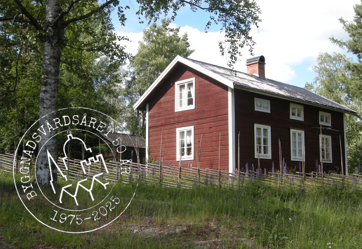 En bild på ett rött äldre timmerhus i skogen med en gärdesgård i förgrunden i gräset.