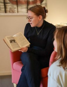 En person lyssnar intresserat på en sagoberättare i en röd fåtölj som visar bild ur en äldre sagobok.