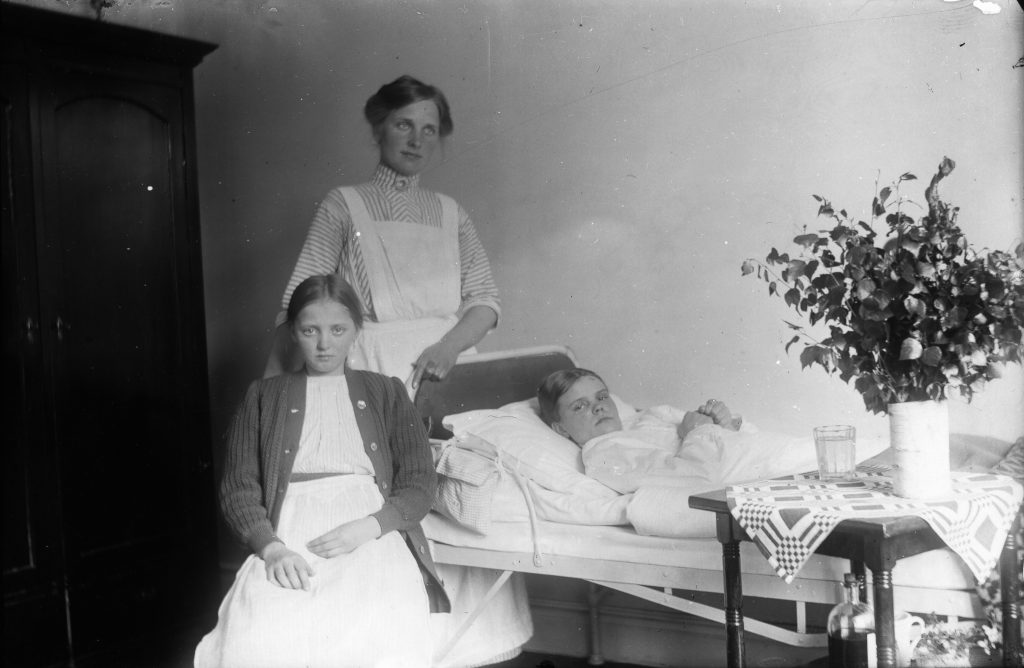 Ett svartvitt foto på en sjuksköterska och ett sjukt barn i en säng och en flicka sittande invid.